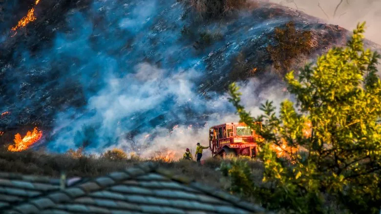 Two firefighters and bulldozer with brushfire burning hillside 