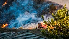 Two firefighters and bulldozer with brushfire burning hillside 